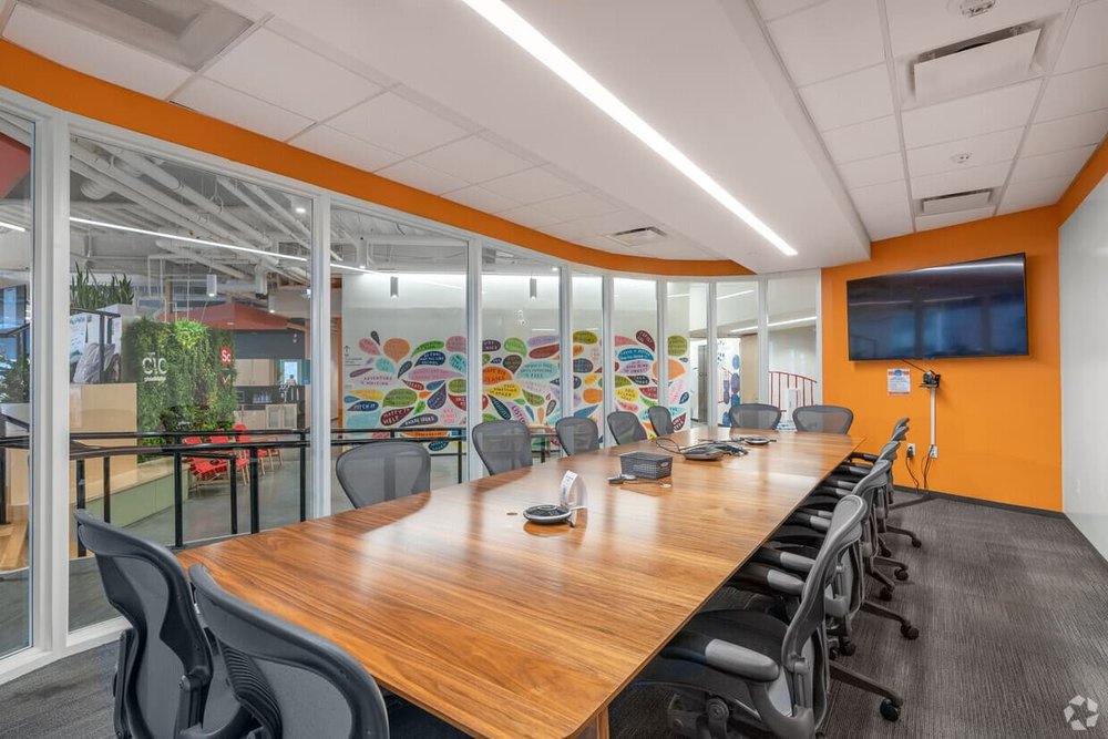 A large conference room with a boardroom table, chairs, video conferencing, mounted TV monitor, whiteboard wall, and glass walls on one side looking out onto a CIC common space.