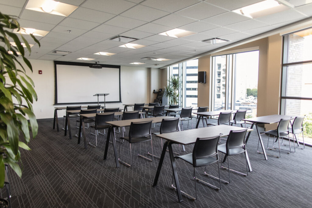Edgewood event space at CIC Providence with tables and chairs in classroom style, projector screen down at the front of the room, podium, and large windows.