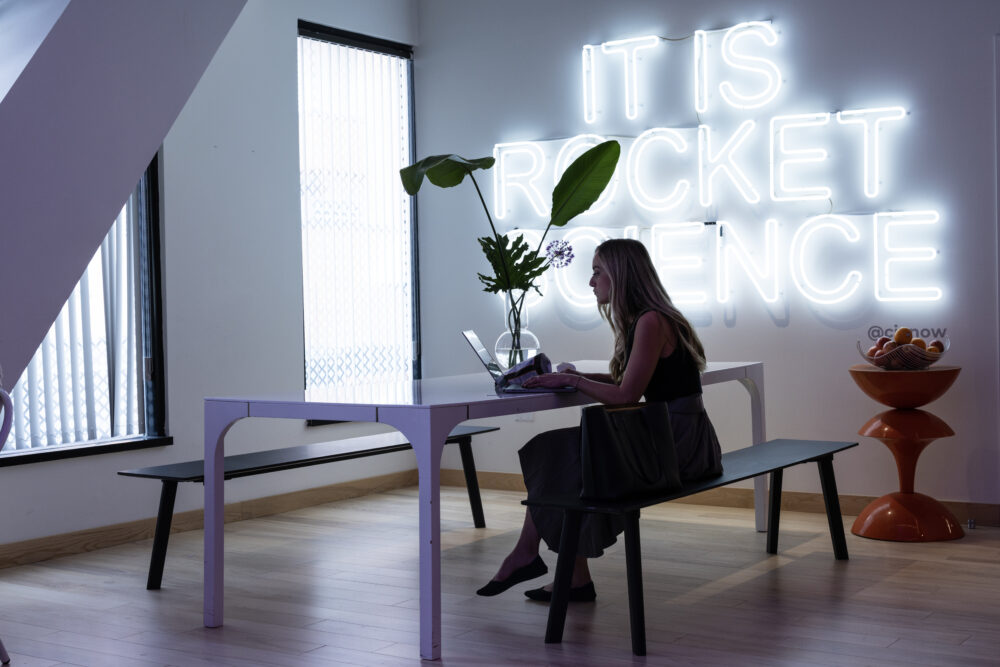 Woman working at a table with a neon sign that reads It Is Rocket Science behind her