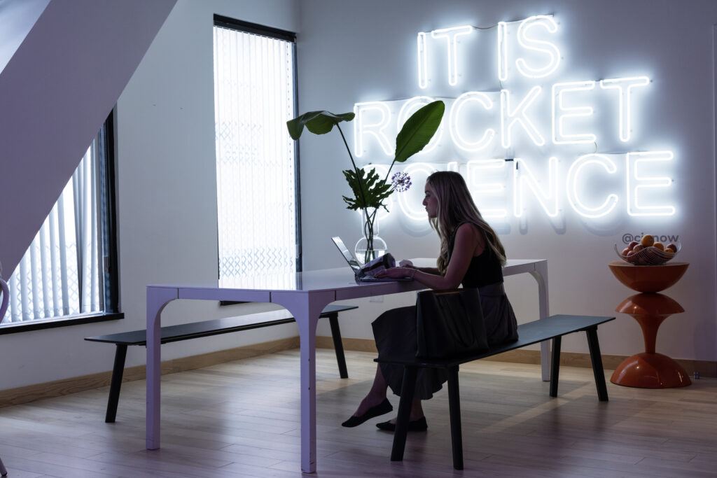 Woman working at a table with a neon sign that reads It Is Rocket Science behind her